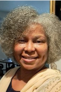 A woman with curly gray hair wearing a tan sweater smiles towards the camera