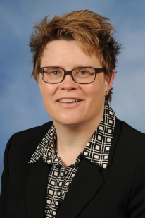 A woman with short brown hair and rectangular glasses wearing a button up and black blazer smiles towards the camera