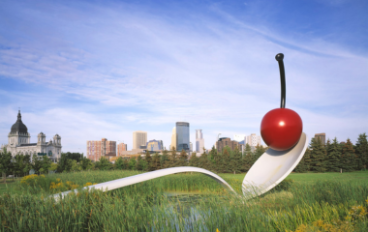 A large white spoon with a red cherry in it amidst a grass field with a city skyline in the background