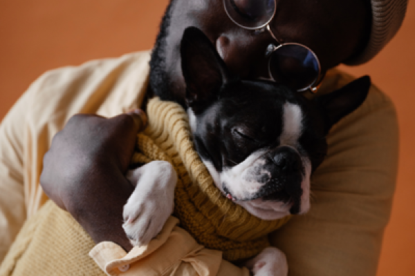 Dark skinned man wearing glasses, beanie, and sweater cradles a sleeping black and white pug